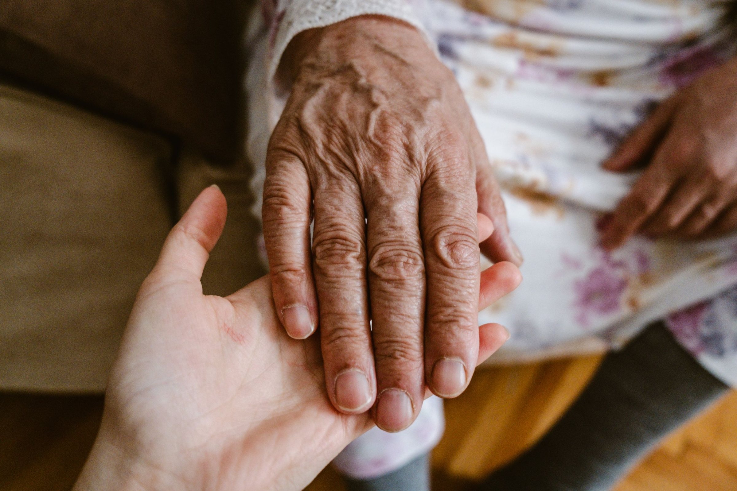 A younger person's hand cradling an older person's hand against the arm of an upholstered chair, with no faces in frame.