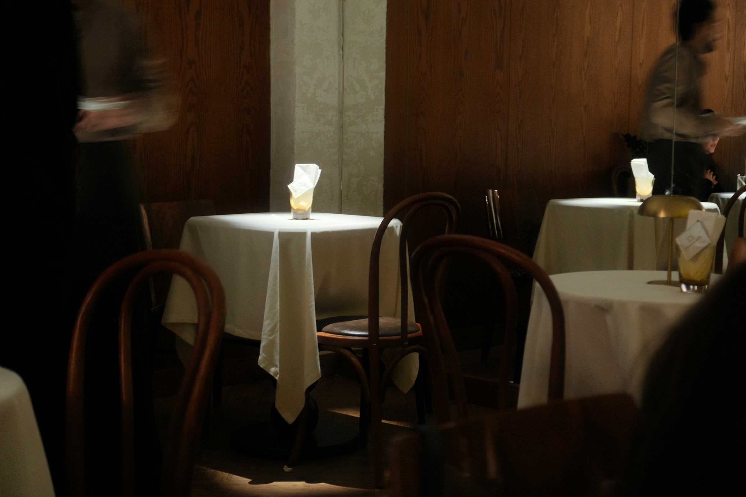 A low-lit dining room with dark wood panelling, bentwood chairs and small round tables laid with white linen, two staff blurred by movement as they cross the room.