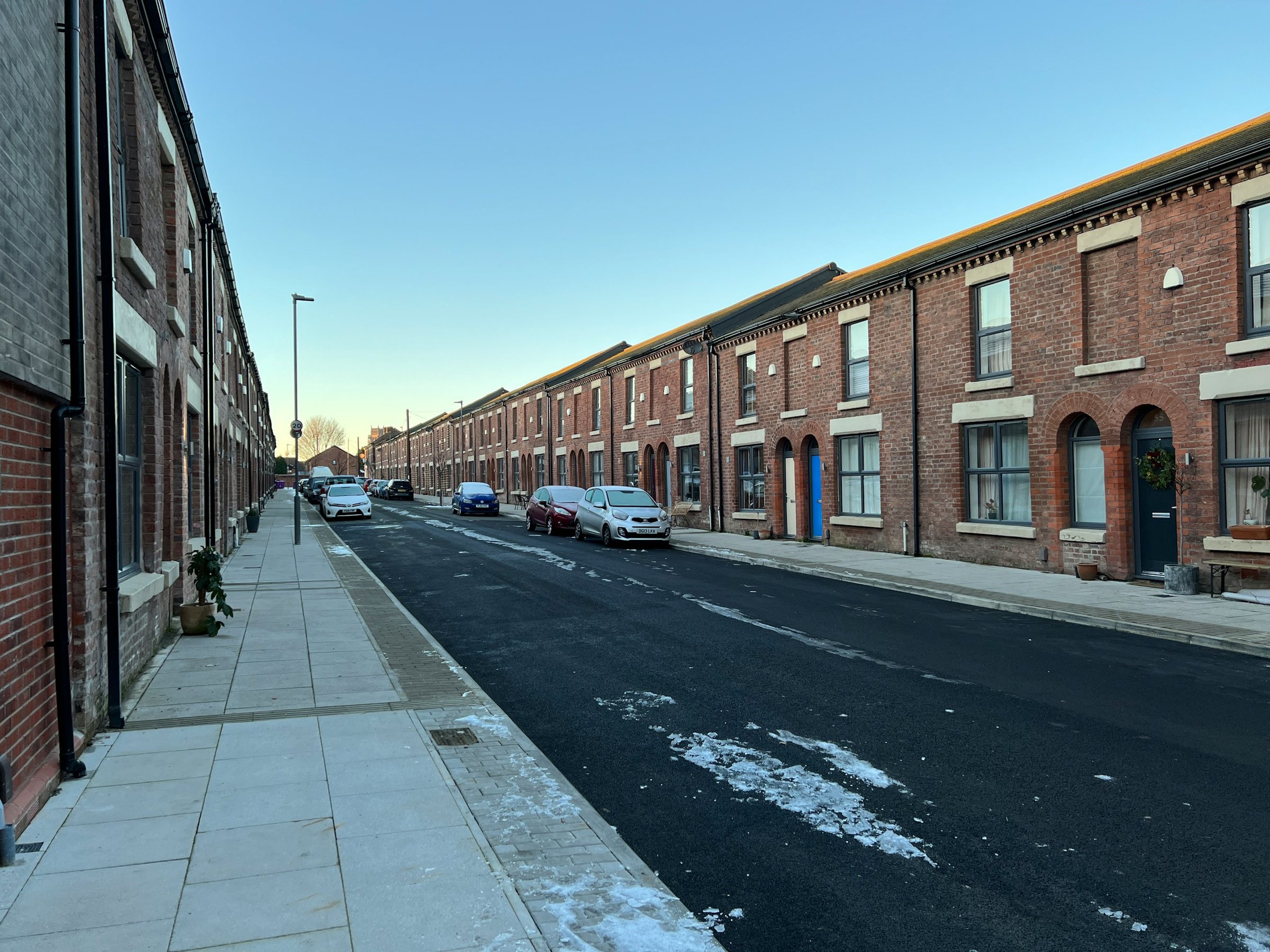 A street of red-brick Victorian terraced houses with arched doorways and sash windows, cars parked along one kerb and patches of melting snow on the road under a pale winter sky.