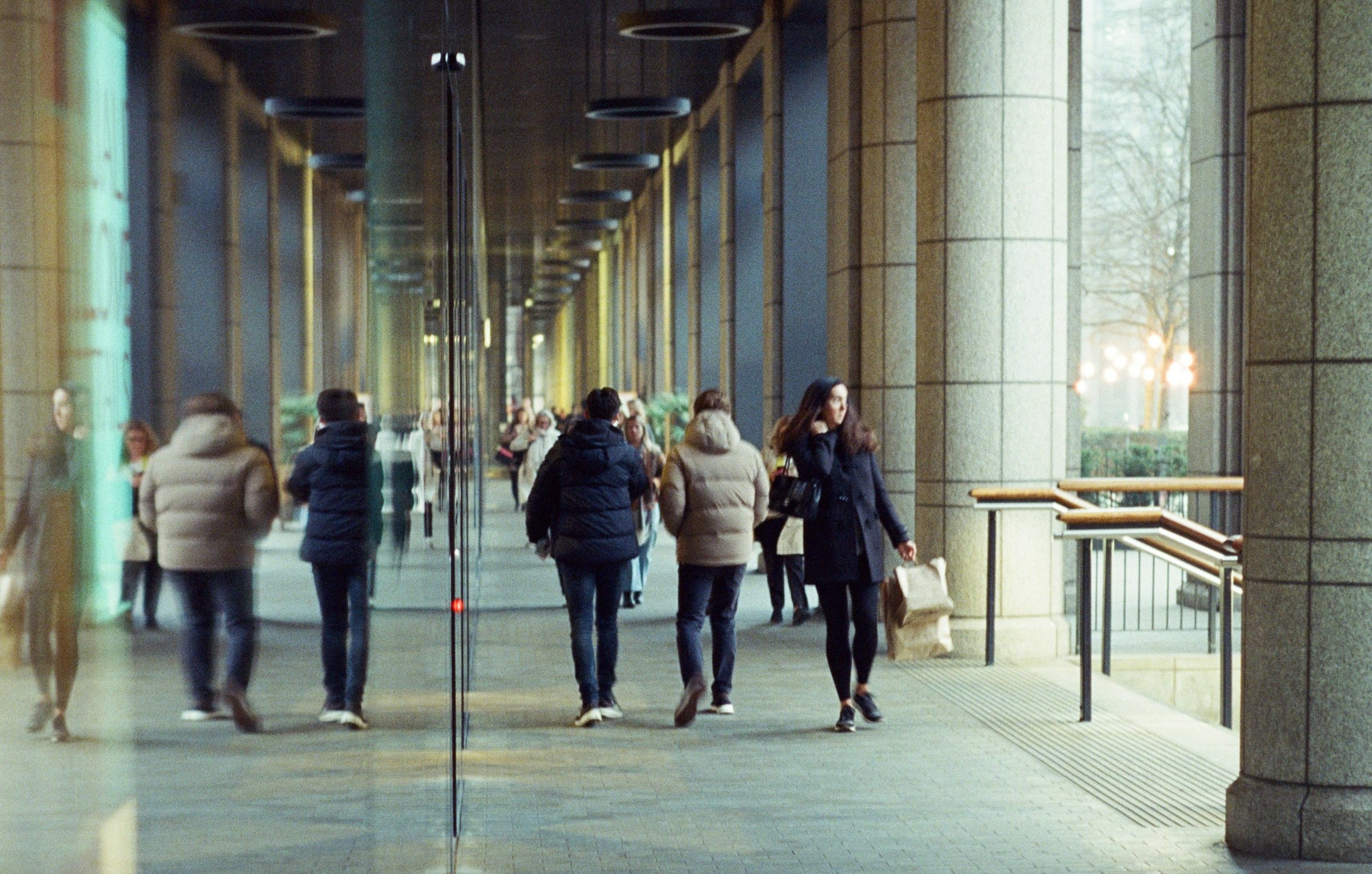 People walking through the daylit stone-and-glass colonnade of a modern building in London, seen from under the canopy on a grey winter afternoon.