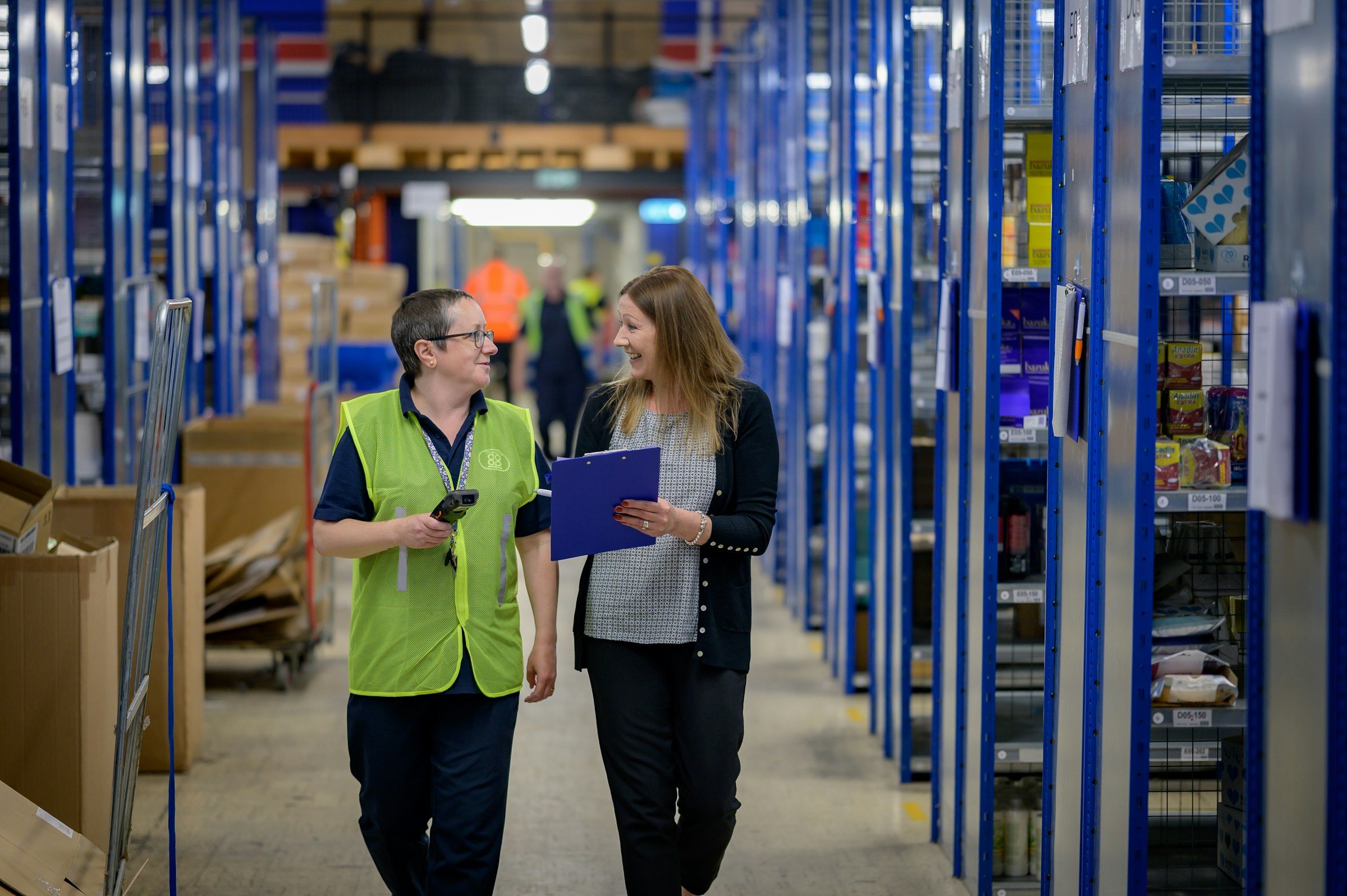 Two warehouse colleagues walk down an aisle of blue steel racking stacked with stock, one in a hi-vis vest carrying a handheld scanner, the other holding a clipboard.